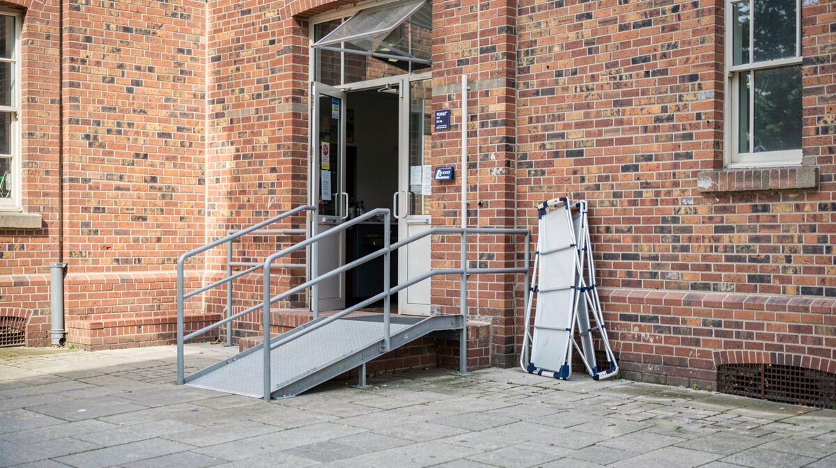 A side entrance of a brick public hall in daylight, a short ramp with metal handrails leading to a propped-open door, a folded portable ramp leaning against the wall beside it, shot from the pavement at standing height.